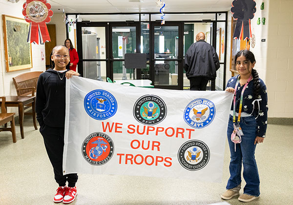 Two students hold a white We Support Our Troops banner with the five U.S. military branch seals in a school hallway.
