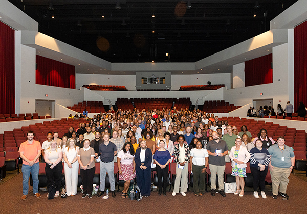 Group photo of hundreds of employees gathered inside a large auditorium during a new employee orientation event, standing together in front of rows of red theater seats.